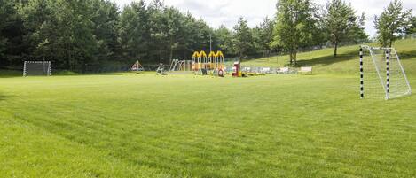 Un terrain herbeux bien entretenu, équipé de deux buts de football, une aire de jeux avec des structures d’escalade jaunes et un espace avec des bancs.