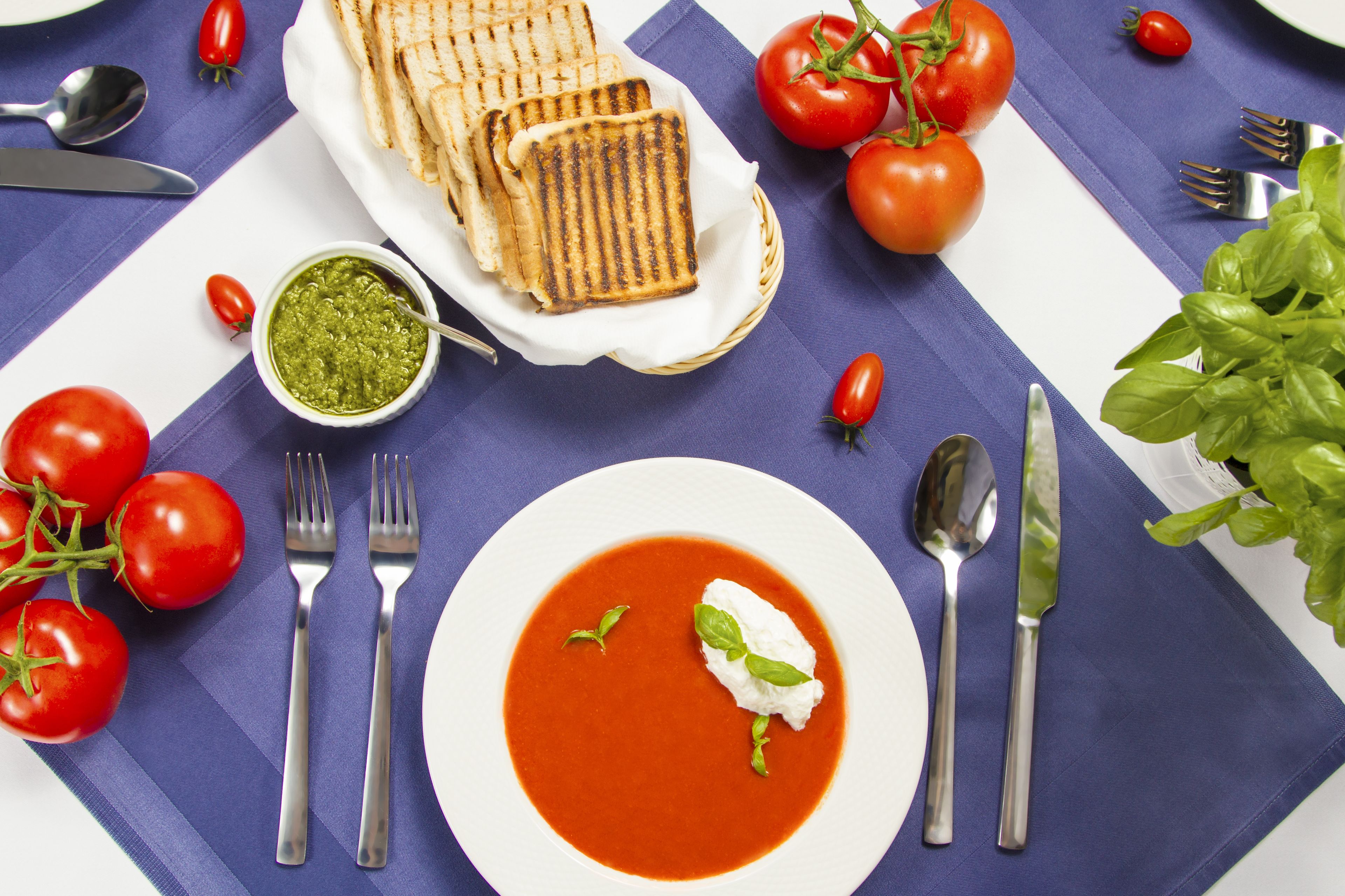 Une table dressée avec une assiette de soupe à la tomate, un bol de pesto, du pain grillé et des tomates fraîches.
