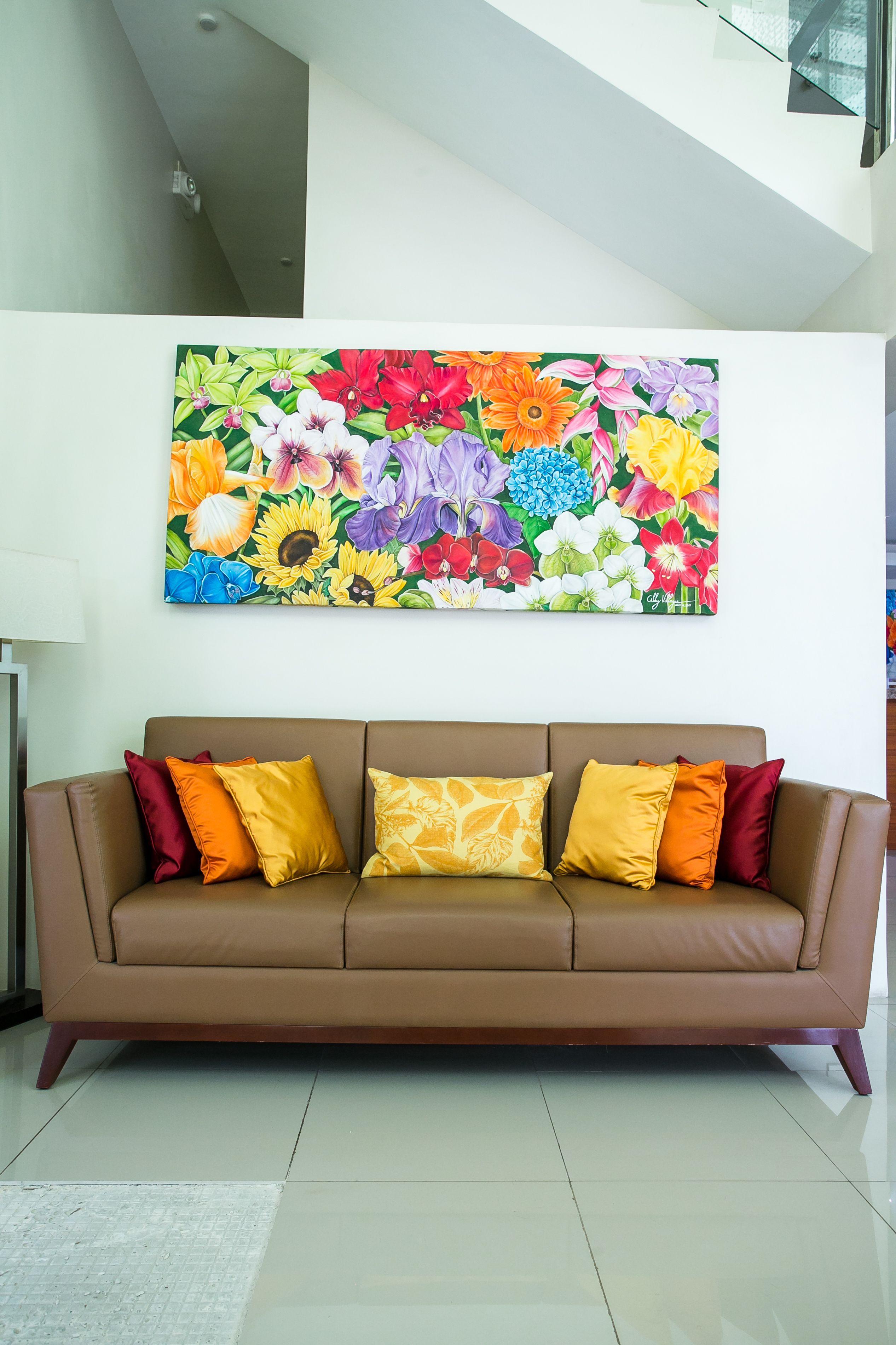 A brown leather sofa with colorful cushions, a large floral painting on the wall, and a modern staircase in the background.