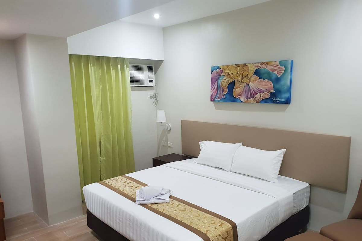 Ein Hotelzimmer mit einem Bett, einem Nachttisch, einer Lampe, einem Gemälde und einem grünen Vorhang.