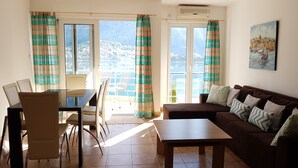 Un salon comprenant un canapé, une table basse, une table à manger et des chaises. De grandes fenêtres sont présentes, ornées de rideaux, ainsi qu’un appareil de climatisation et un tableau accroché au mur.