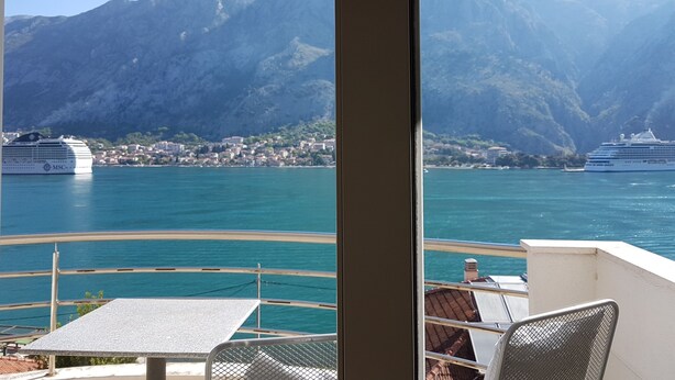 Un balcon avec une table et des chaises donnant sur une étendue d’eau où l’on peut apercevoir des navires de croisière, avec un paysage montagneux en arrière-plan.