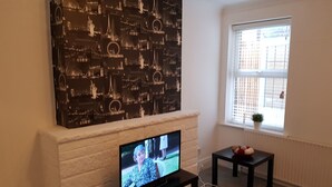Un soggiorno con televisione a schermo piatto su un mobile, un camino, un grande quadro con una veduta della città e una finestra con persiane.