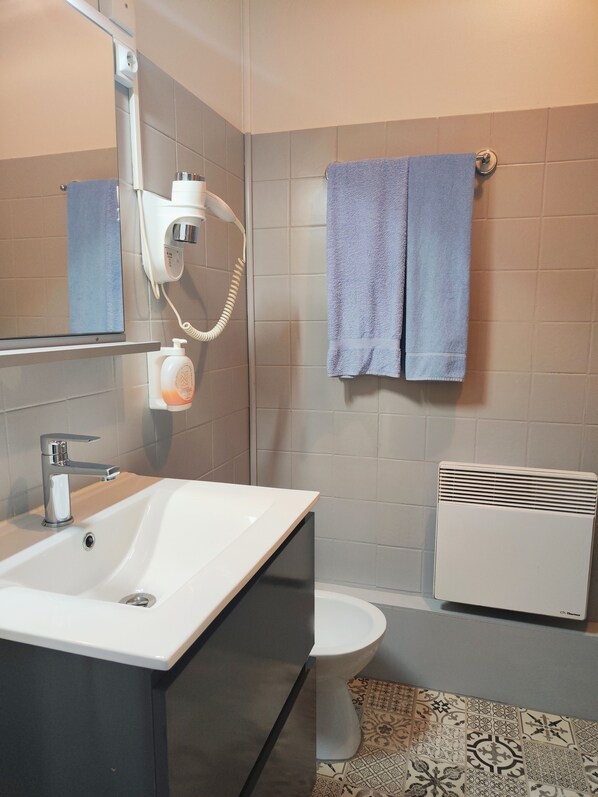 Un baño con lavamanos blanco, un espejo, un secador de pelo, un perchero con dos toallas, un inodoro y un radiador.