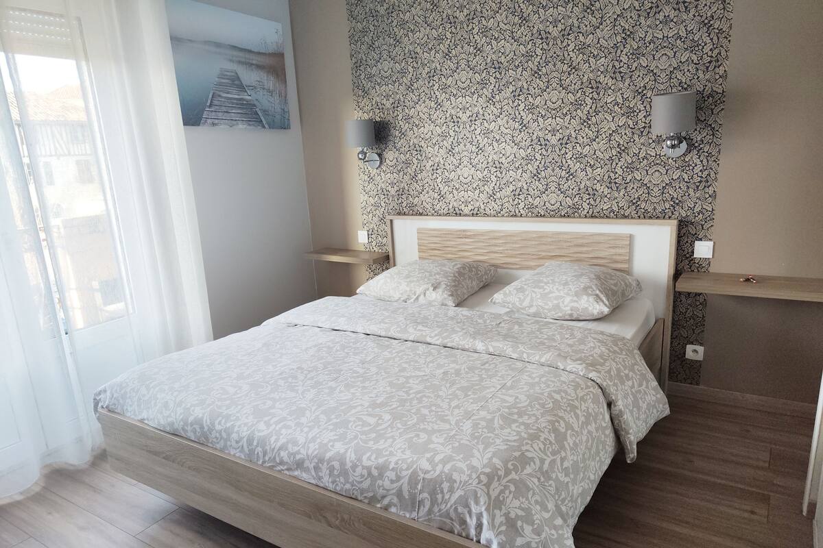 Un dormitorio con cama, cabecera, dos mesitas de noche, lámpara de pared y papel tapiz estampado.