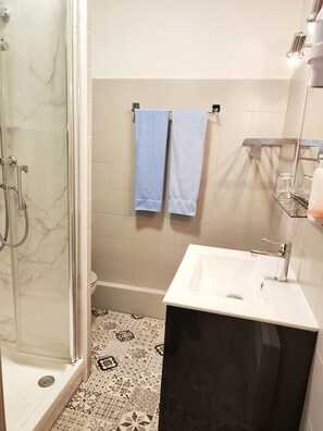 A bathroom with a glass-enclosed shower, a white sink, and a tiled floor with a patterned design. - Hôtel Restaurant Le Commerce (Nogaro)
