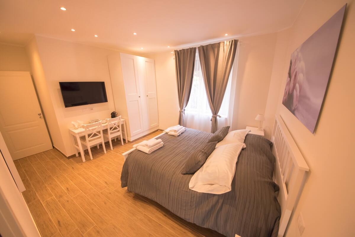 A hotel room with a large bed, a TV mounted on the wall, a desk with a chair, and a window with curtains.