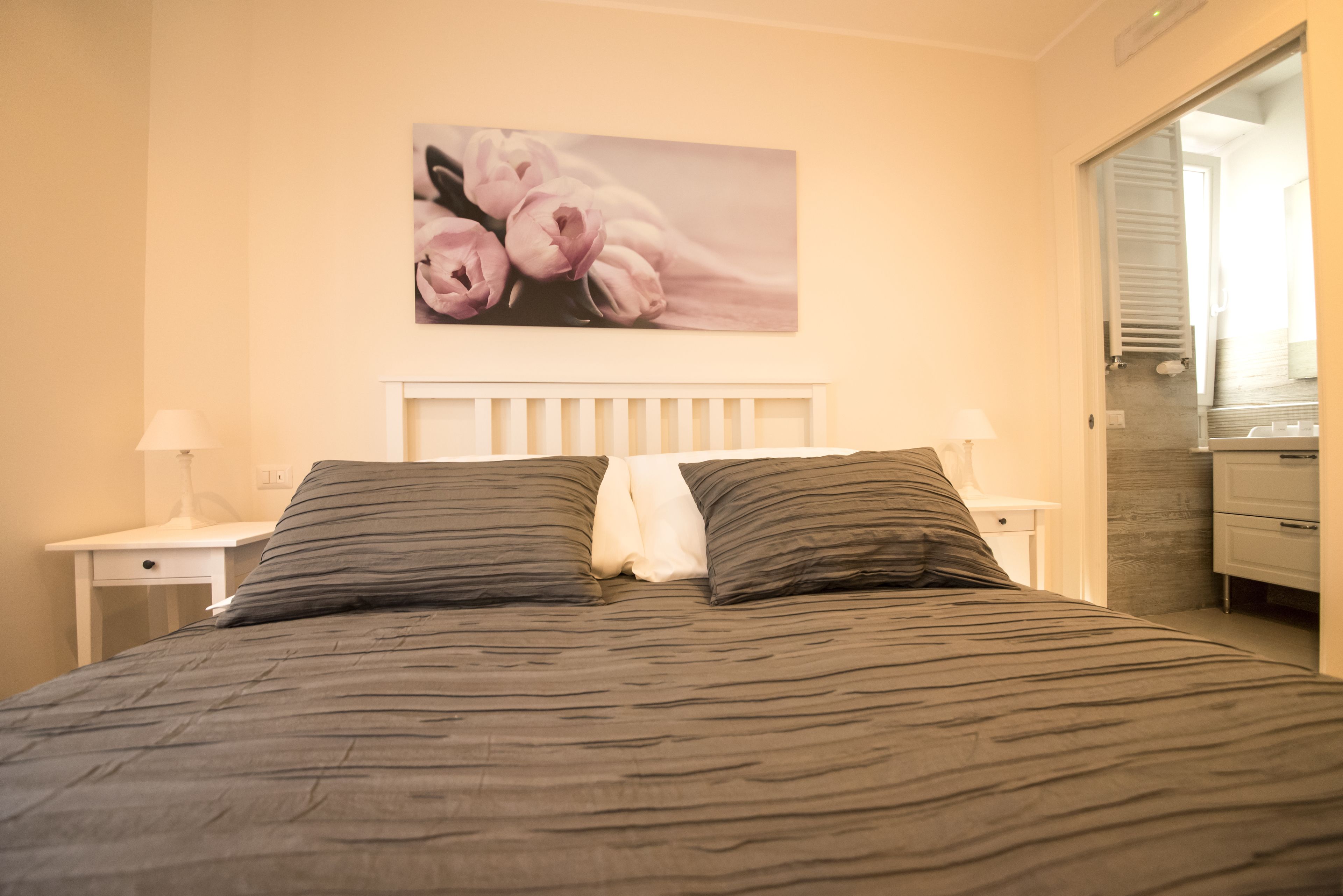 A neatly made bed with a striped bedspread, two bedside lamps, a white nightstand, and a wall-mounted picture of pink flowers.