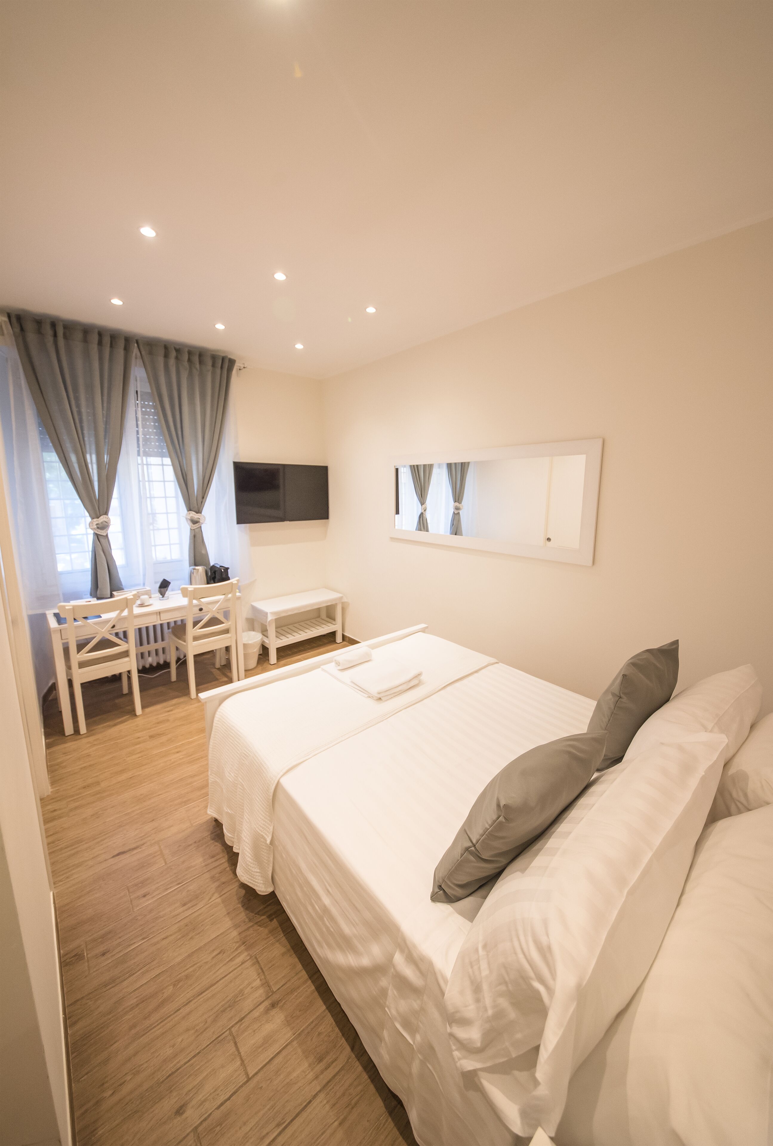 A hotel room with a bed, a desk, a chair, a TV mounted on the wall, and a window with curtains.
