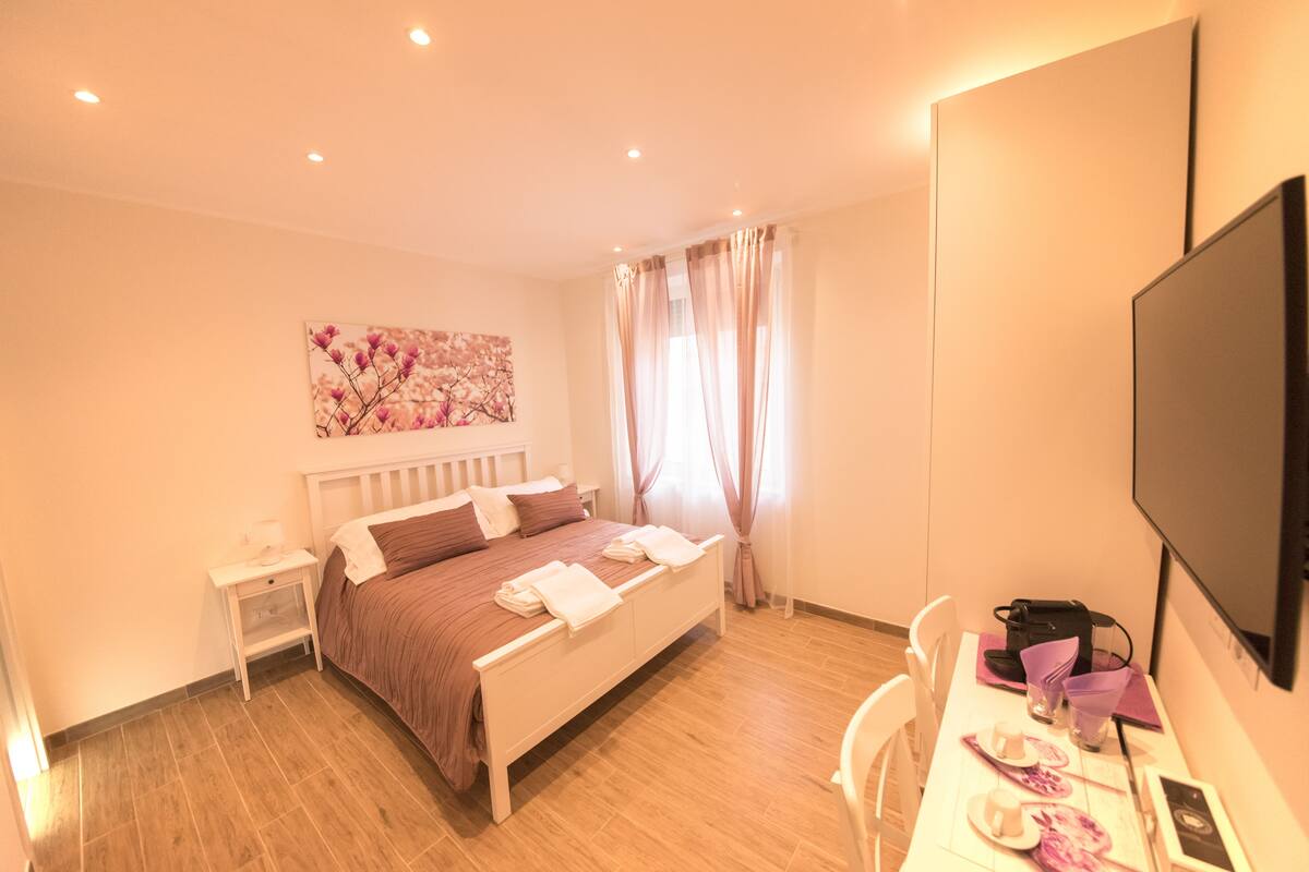 A bedroom with a bed, bedside tables, a desk with a chair, a flat-screen TV, and a window with curtains.
