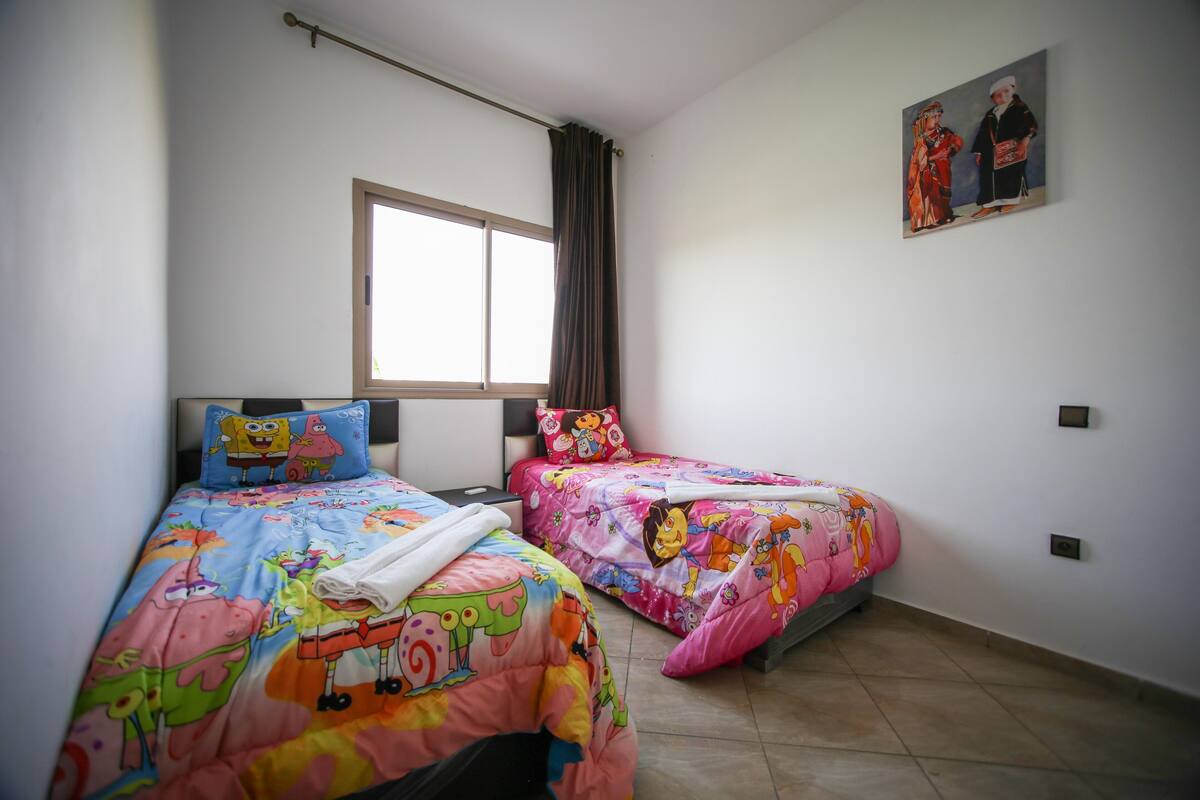 A bedroom with two beds, each with colorful bedding and cartoon character prints. There is a window with a curtain, a painting on the wall, and a ceiling light fixture.