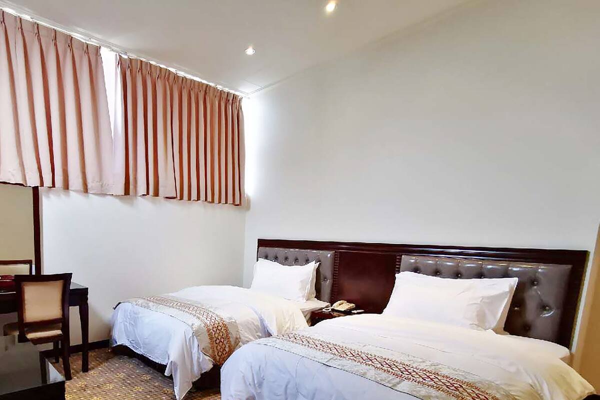 Ein Hotelzimmer mit zwei Einzelbetten, einem Schreibtisch mit Stuhl und einem großen Fenster mit Vorhängen.
