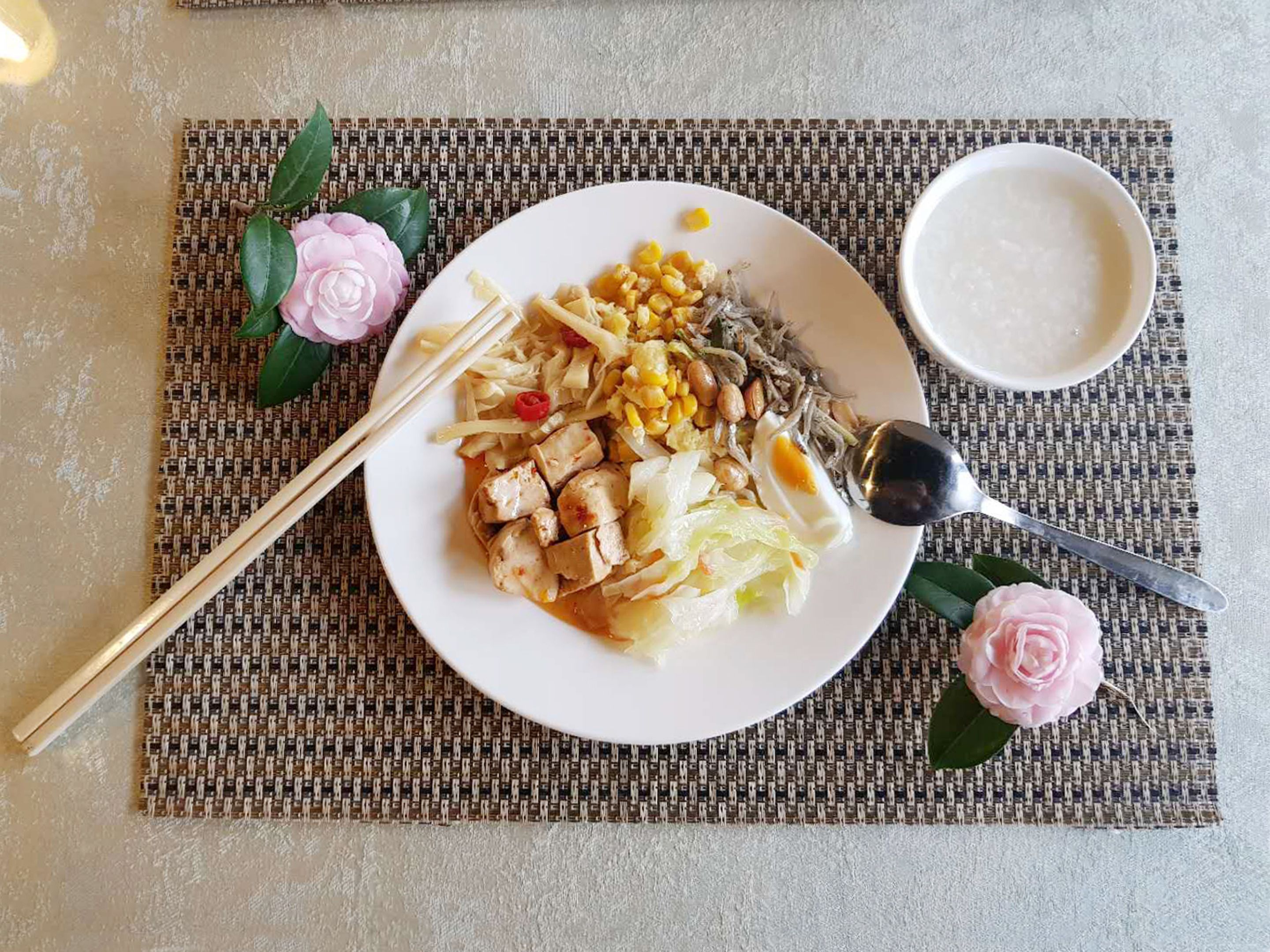 A plate of food with chopsticks and a spoon, a bowl of soup, and decorative flowers on a woven placemat.