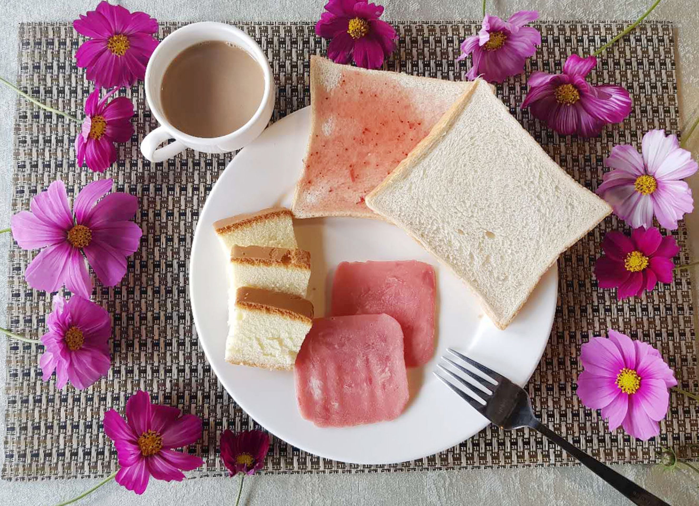 Une assiette contenant des tranches de pain blanc, de la confiture rose et de la gelée rose, accompagnée d’une tasse de café et d’une fourchette, le tout posé sur un set de table en fibres tressées orné de fleurs roses.