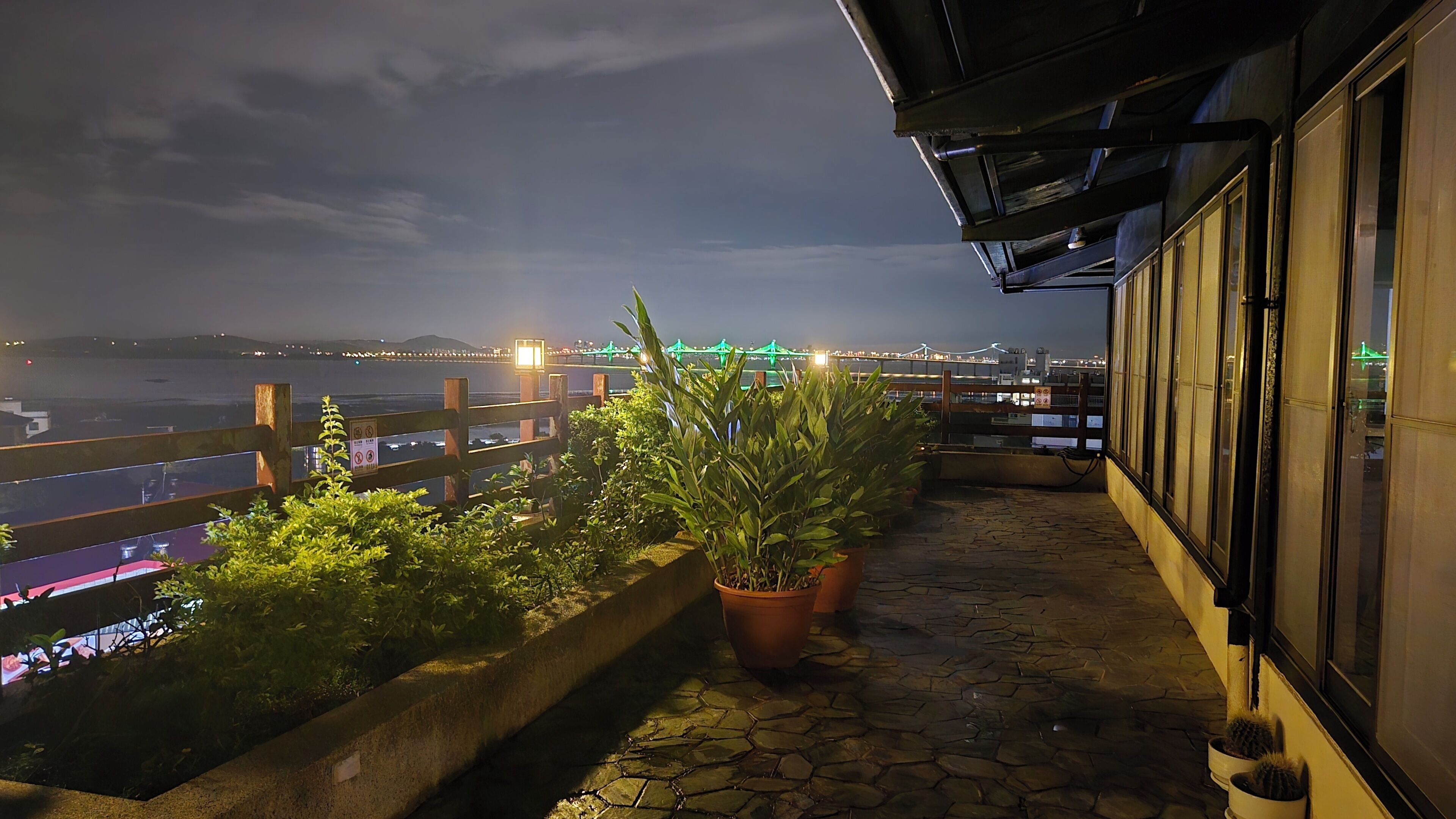 A balcony with potted plants, a stone floor, and a view of a bridge and city lights.