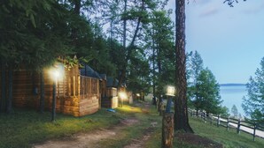 Une cabane en bois, accessible par un sentier de terre, entourée d’arbres et offrant une vue sur une étendue d’eau.