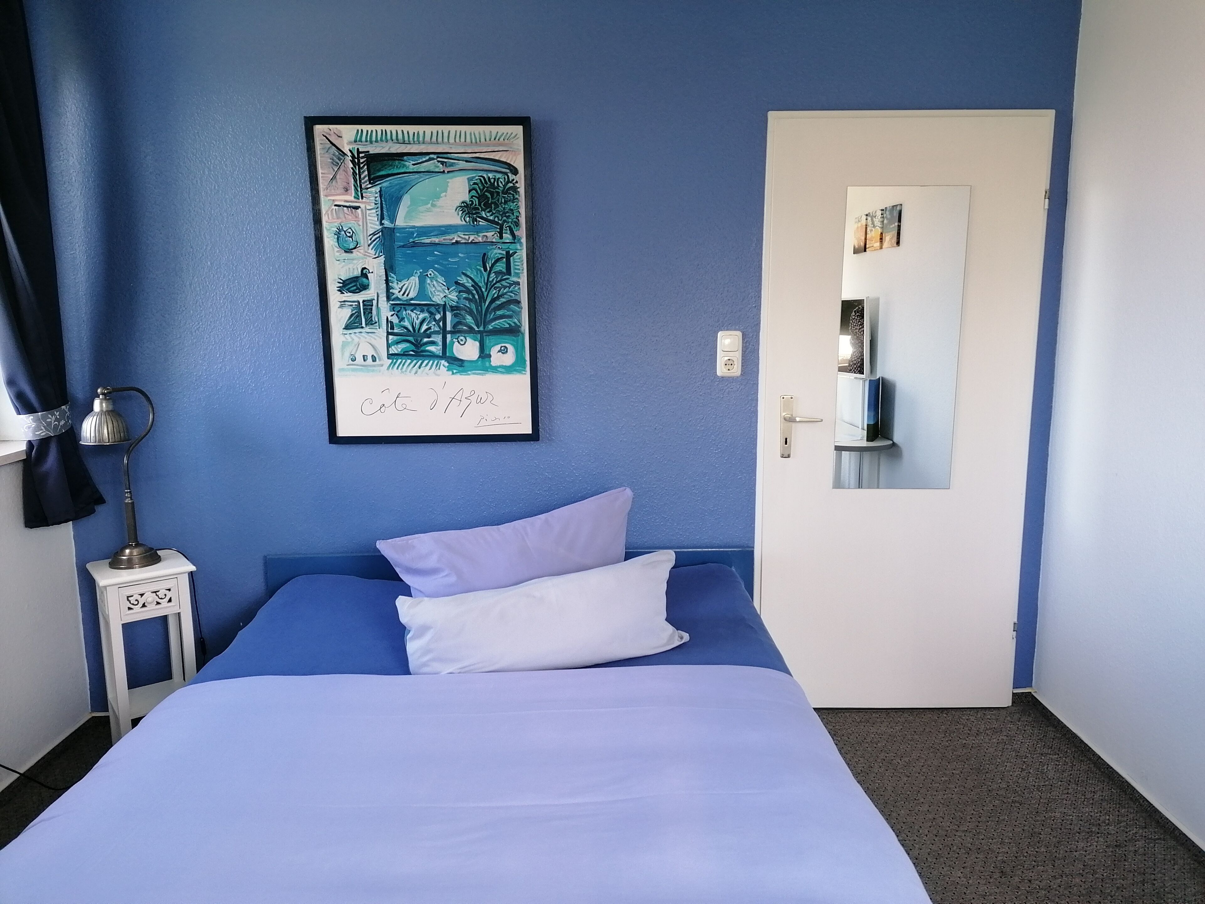Une pièce comprenant un lit, une table de chevet, une lampe, une porte et un mur orné d’un tableau encadré.