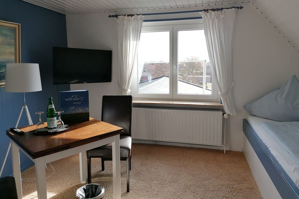 A hotel room with a wooden table, a chair, a TV mounted on the wall, a window with curtains, and a bed with a striped pillow.