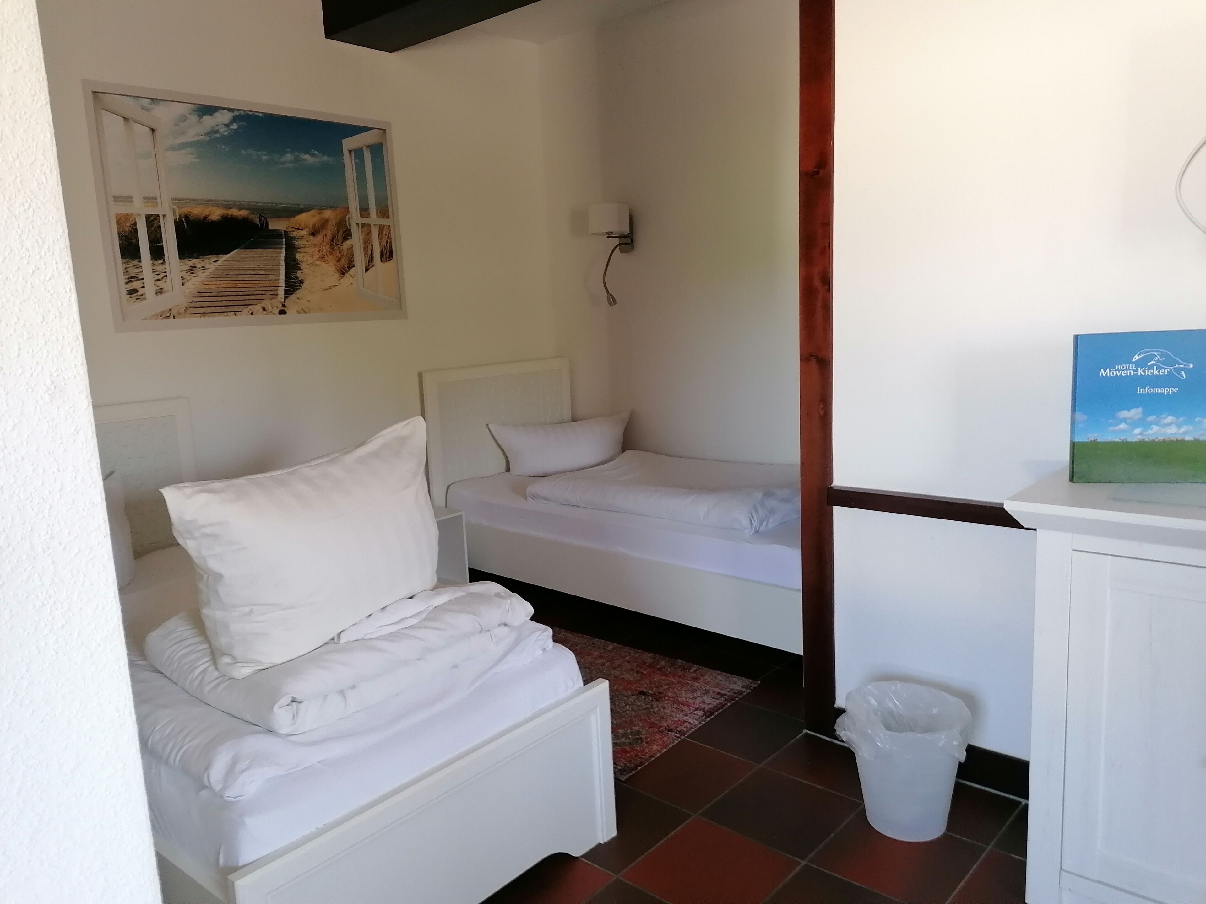 A small, clean room with two single beds, a white wall, a painting of a beach, and a trash bin.