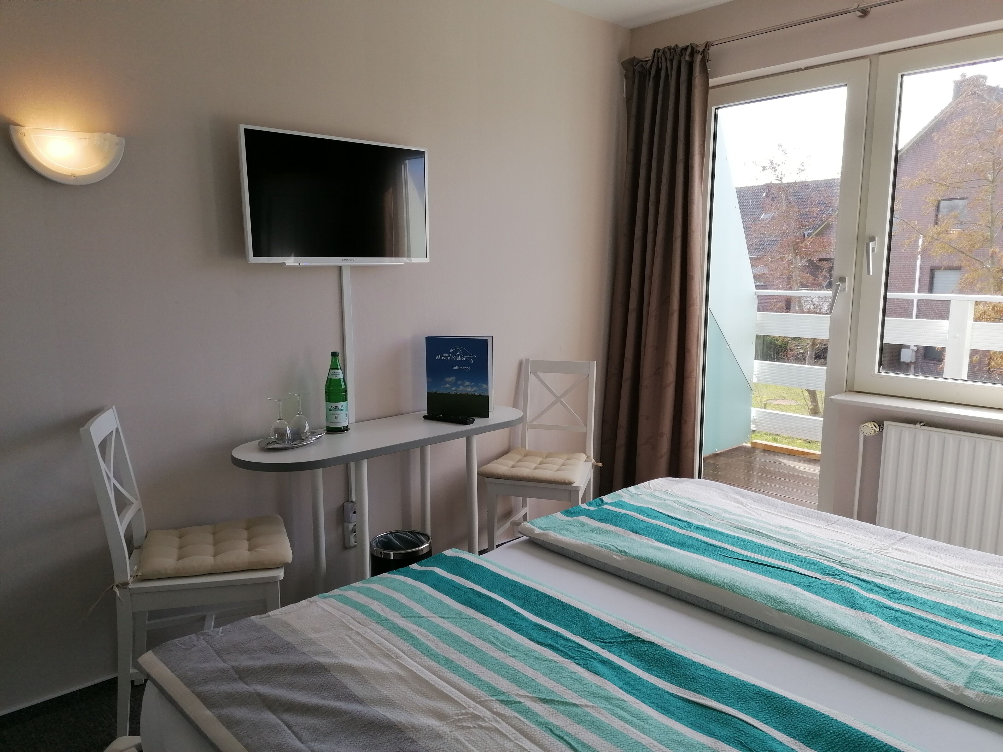 A hotel room with a bed, a chair, a small table with a bottle and glasses, a TV mounted on the wall, and a window with curtains.