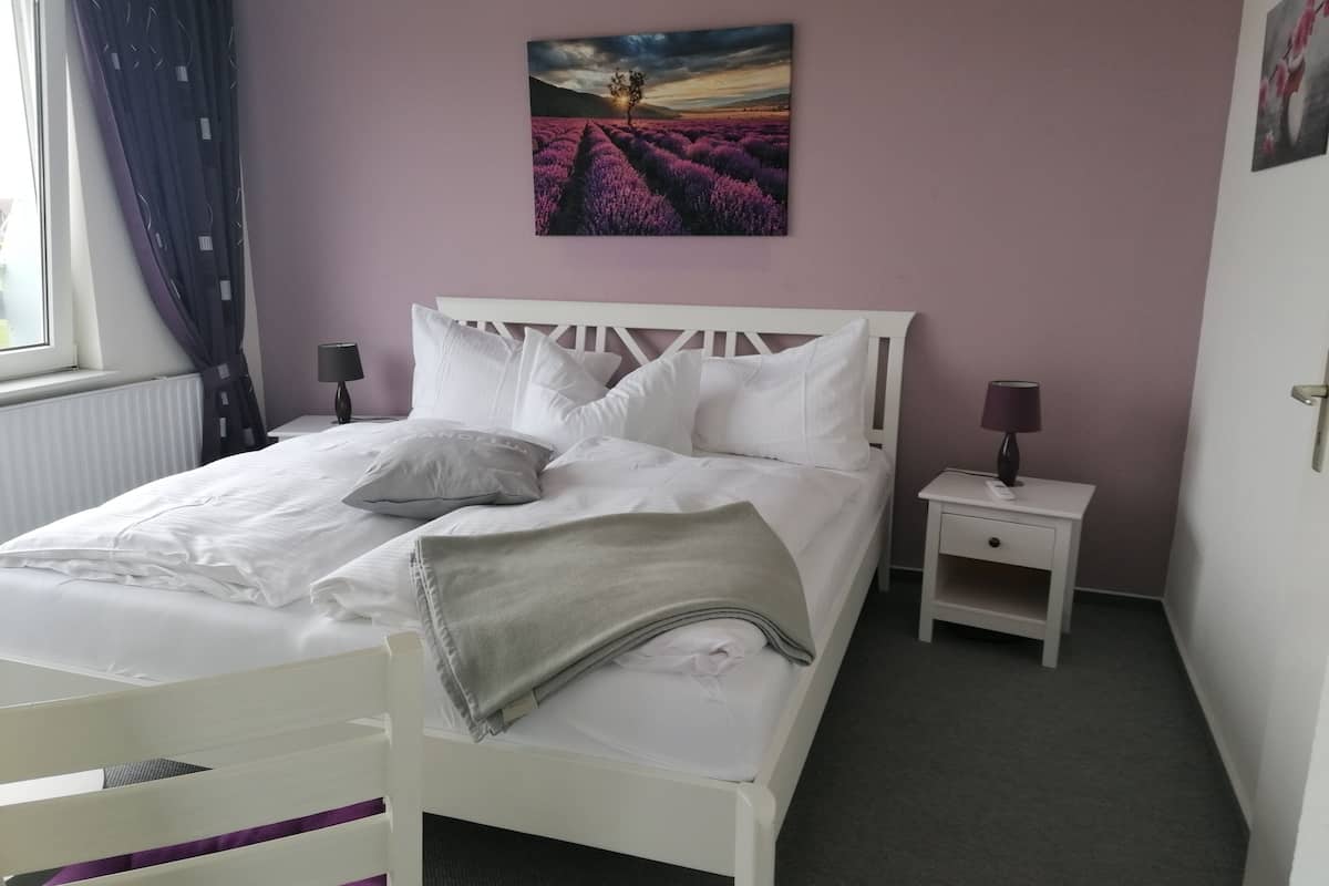 A neatly made bed with white linens and pillows, a small bedside table with a lamp, and a painting of a lavender field on the wall.
