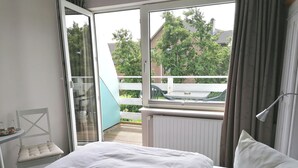 A bedroom with a large bed, a glass door leading to a balcony, and a view of a garden and buildings outside. - Frühstückshotel Möven Kieker (Friedrichskoog)