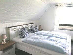 Un dormitorio con techo inclinado, una cama con ropa de cama blanca y almohadas a rayas, una mesita de noche con una lámpara y una ventana con cortinas.