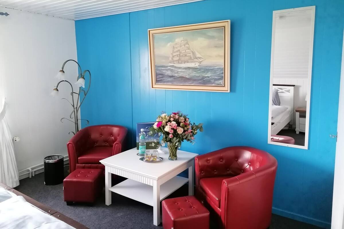 A room with a blue wall, a framed painting of a ship, a white table, and two red chairs.