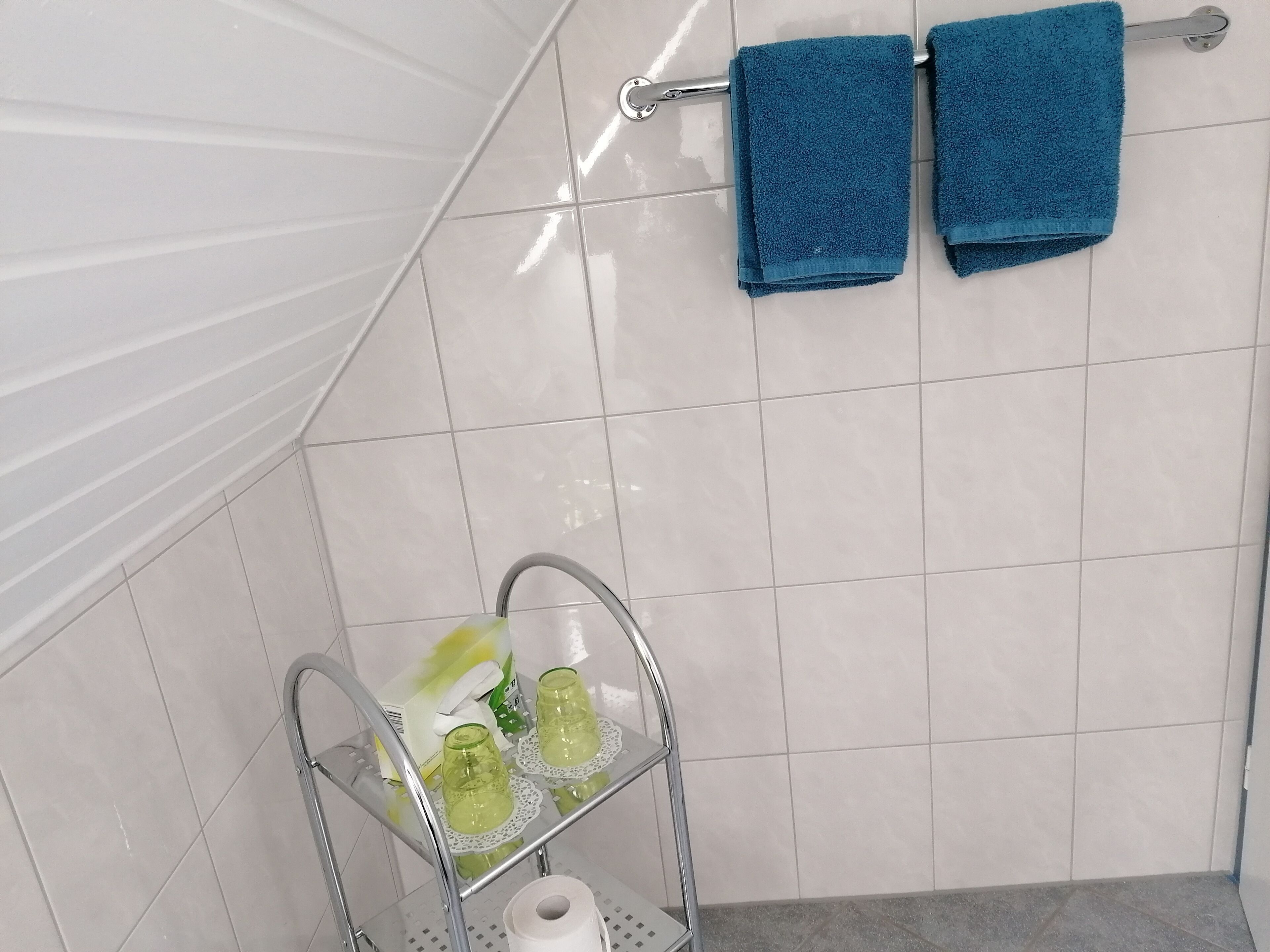 A bathroom with white tiles, a towel rack with two blue towels, and a glass shelf holding two green glass bottles and a roll of toilet paper.