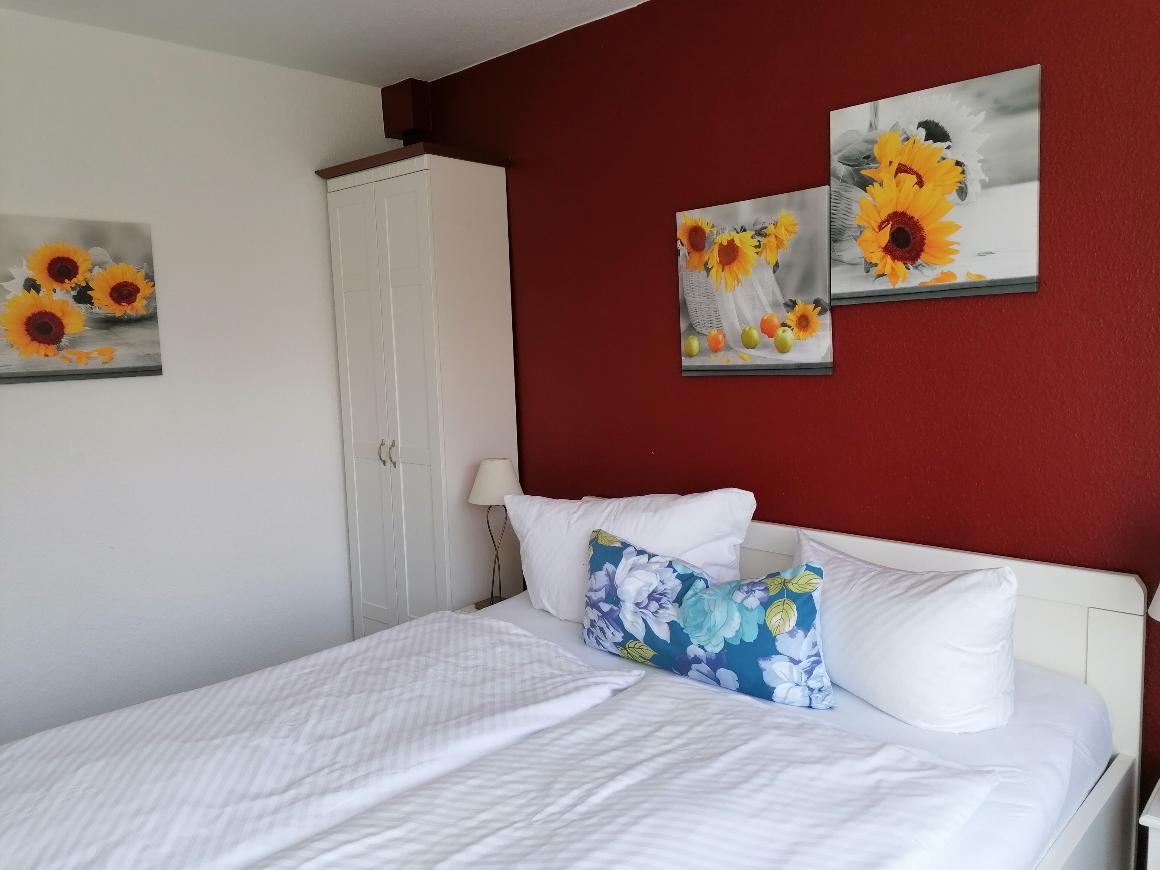 Un dormitorio con una cama, paredes blancas, una pared de acento roja y dos cuadros de girasoles.