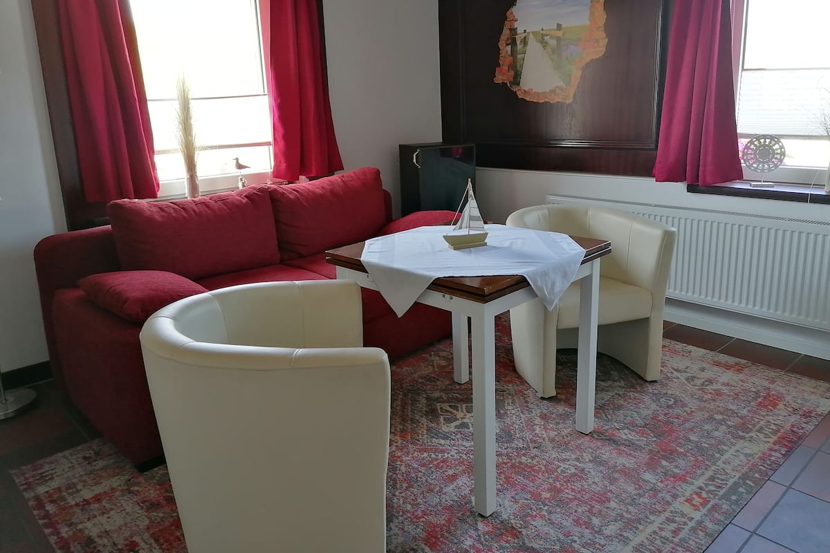 A room with a red sofa, a white armchair, a small table with a white tablecloth, and a large window with red curtains.
