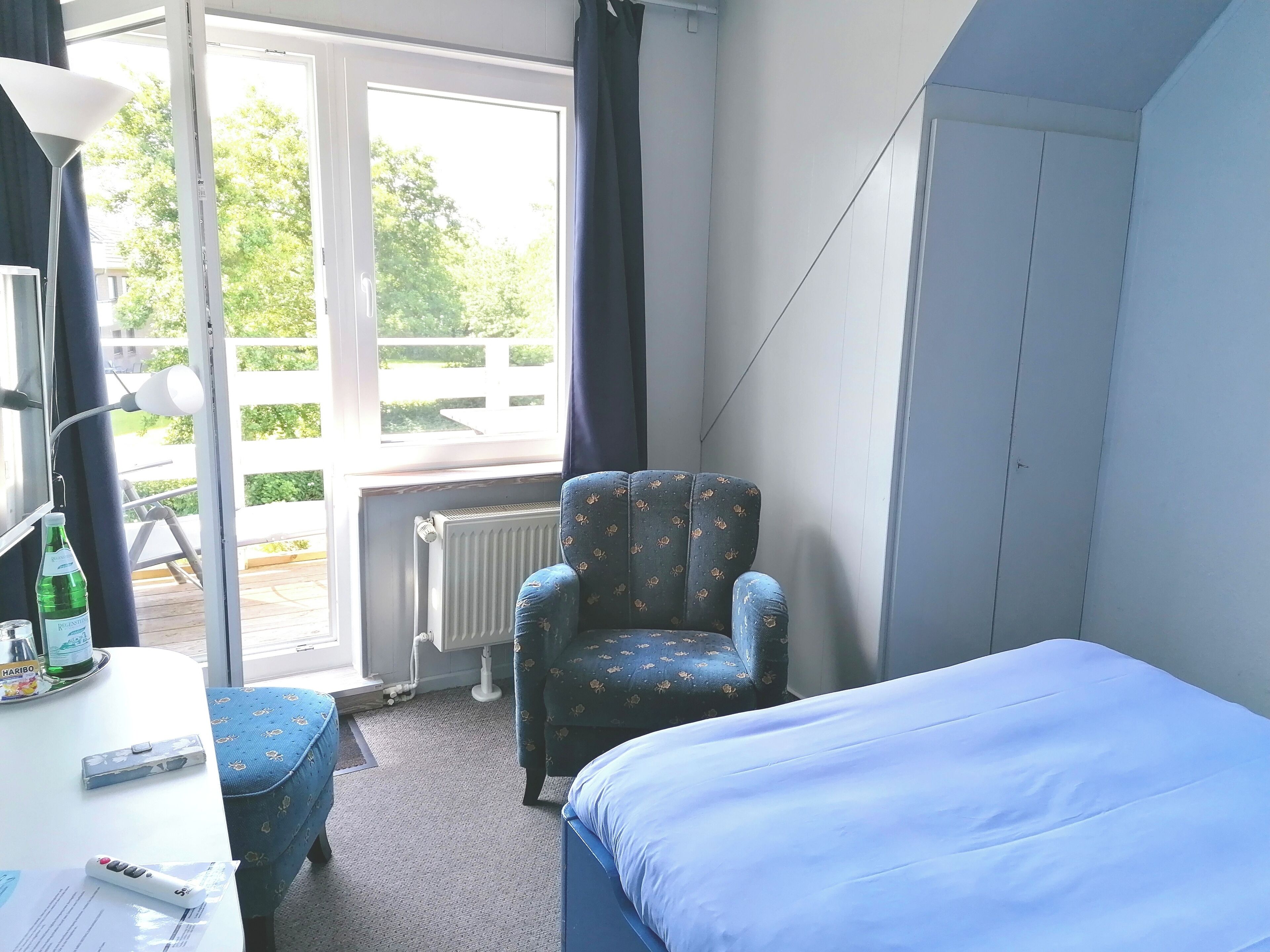 Ein Hotelzimmer mit einem Bett, einem Sessel, einem Schreibtisch mit Lampe, einem Heizkörper und einem Fenster mit Vorhängen.