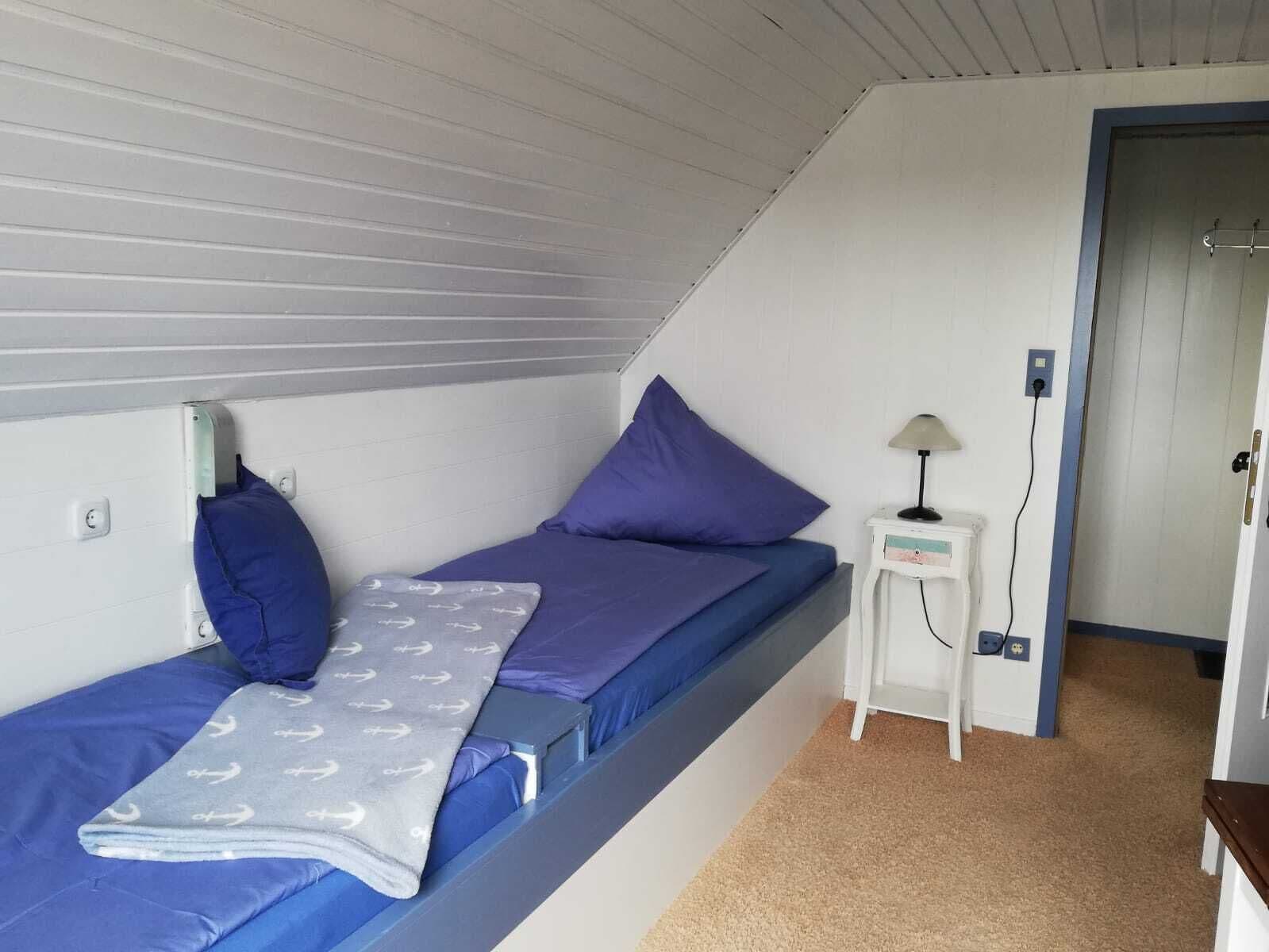 A small, single-bed room with a sloped ceiling, a bedside table with a lamp, and a power outlet.