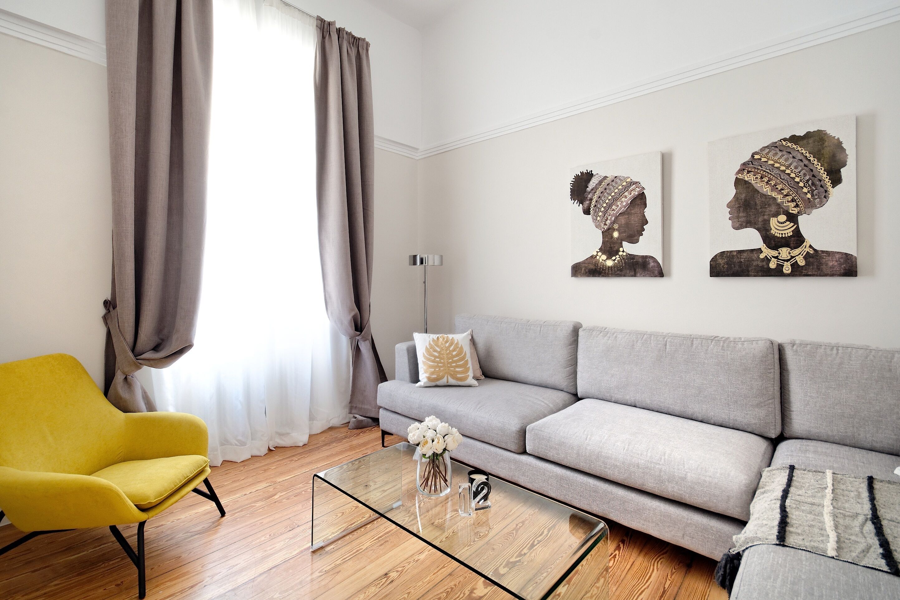 A modern living room with a yellow armchair, a grey sofa, and a glass coffee table. There are two wall art pieces depicting stylized human profiles.