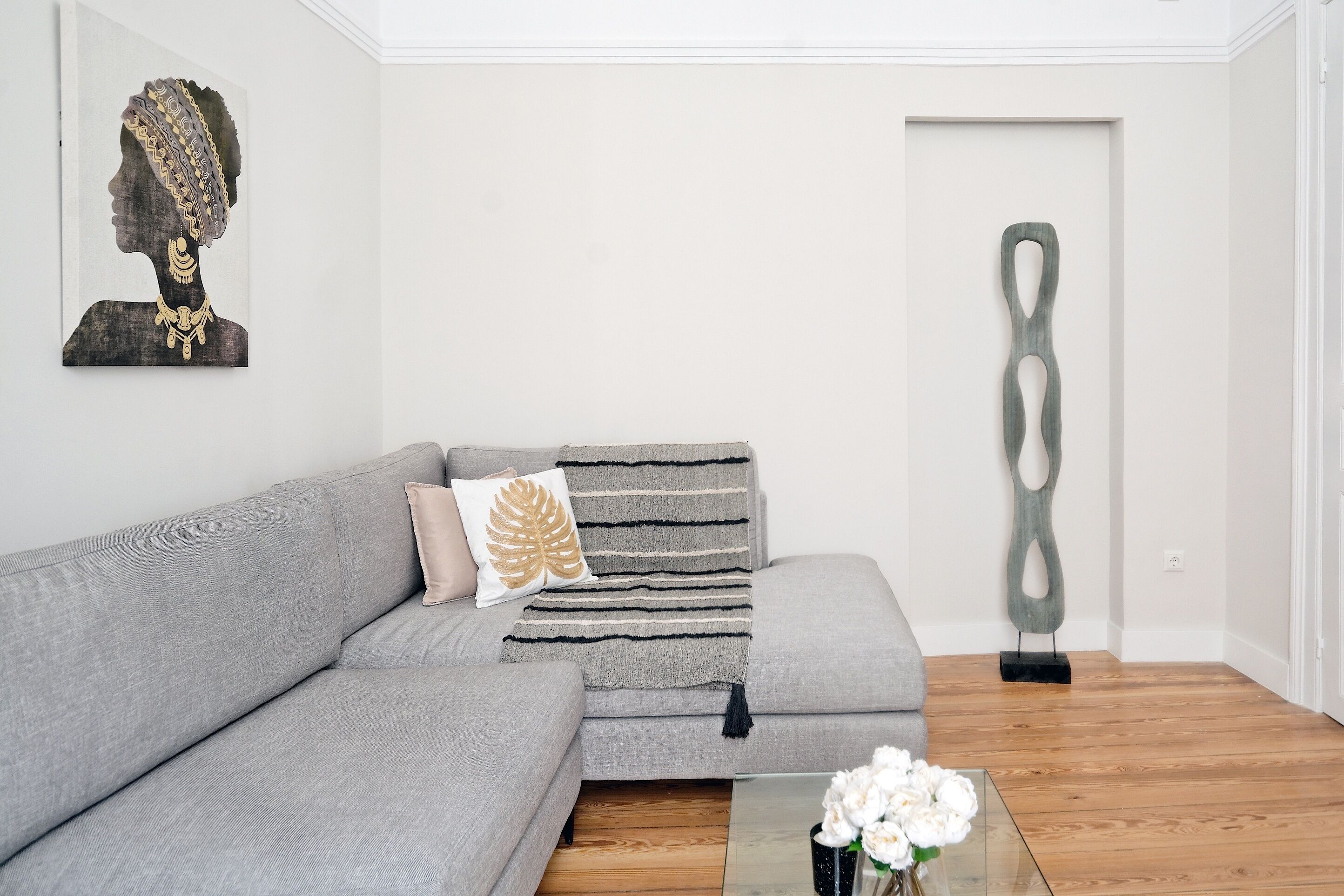 A modern living room with a grey sofa, a decorative pillow, a striped throw blanket, a minimalist sculpture, and a glass coffee table with white flowers.