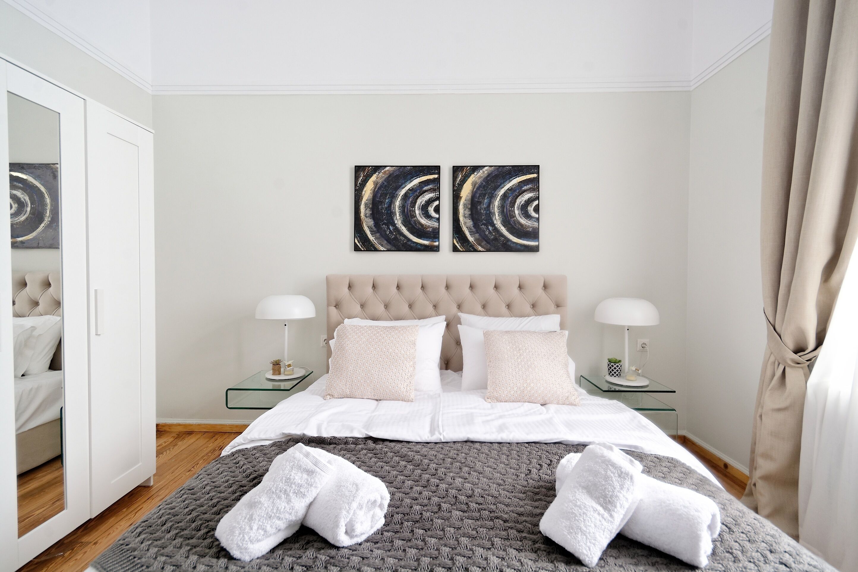 A neatly made bed with white linens and two pillows, a glass bedside table with a lamp, and two abstract paintings on the wall.