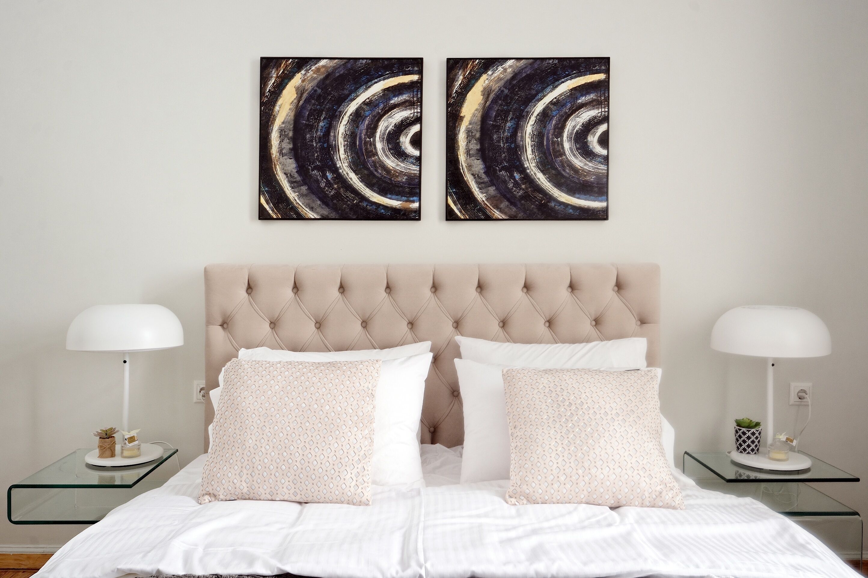 A bedroom with a tufted headboard, two bedside tables with lamps, and two abstract wall art pieces.