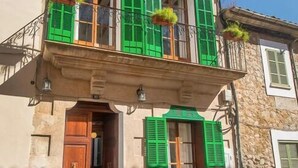 Un edificio de dos pisos con contraventanas verdes, un balcón con plantas colgantes y fachada de piedra.
