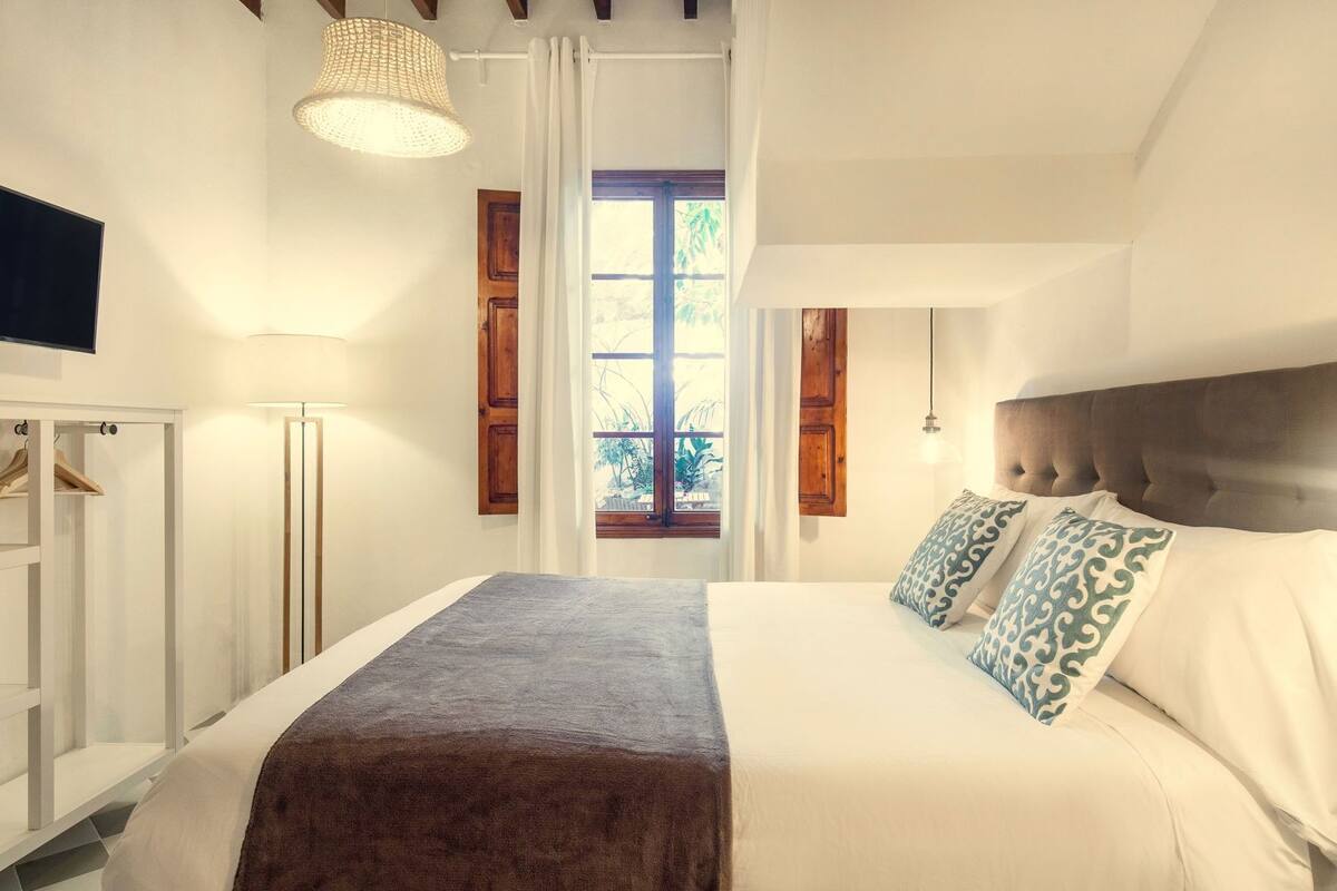 Un dormitorio con cama, armario, lámpara colgante y ventana con cortinas.