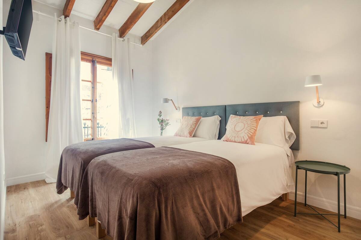 Un dormitorio con dos camas, suelo de madera, una mesita y una ventana con cortinas.