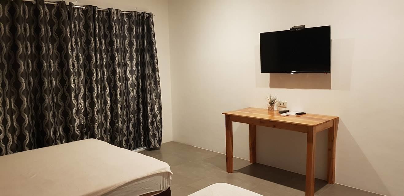 A small, simple room with a bed, a wooden table, a TV mounted on the wall, and a patterned curtain.
