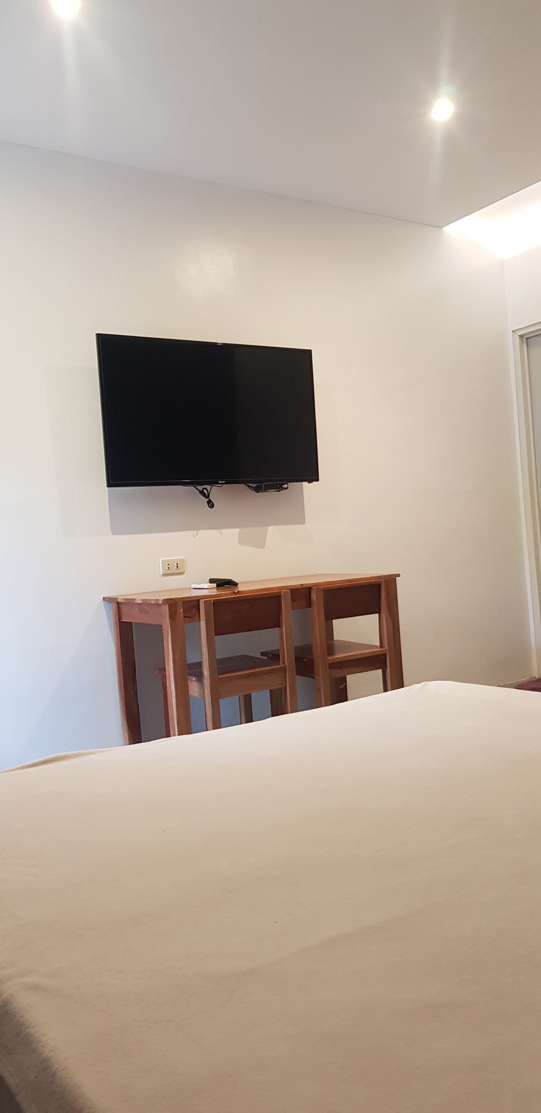 A bedroom with a bed, a wooden nightstand, a flat-screen TV mounted on the wall, and a door to the right.