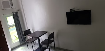 Quarto com mesa, duas cadeiras, televisão na parede e janela com cortinas.