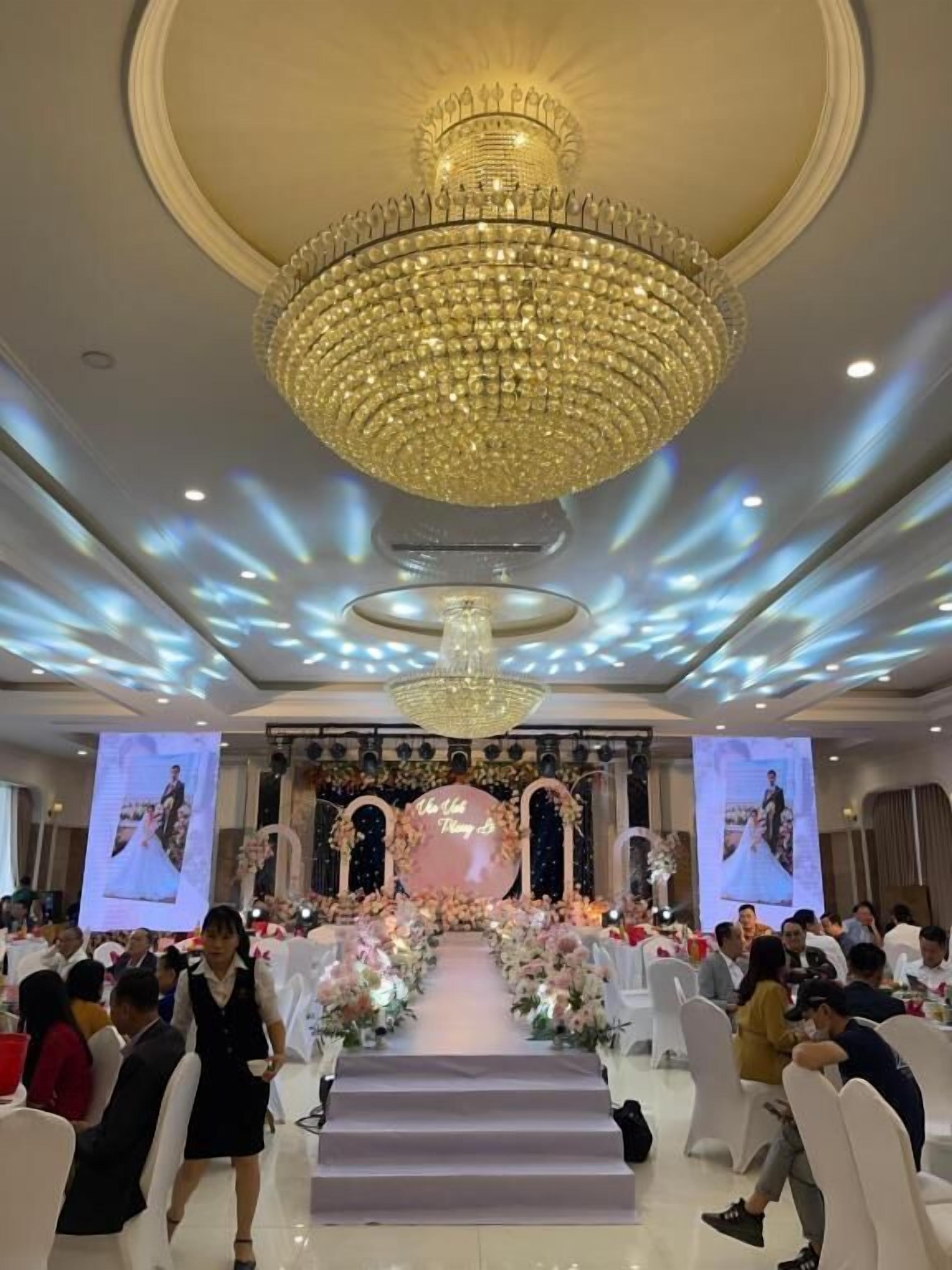 A formal event with a stage, attendees seated on white chairs, and a large chandelier.
