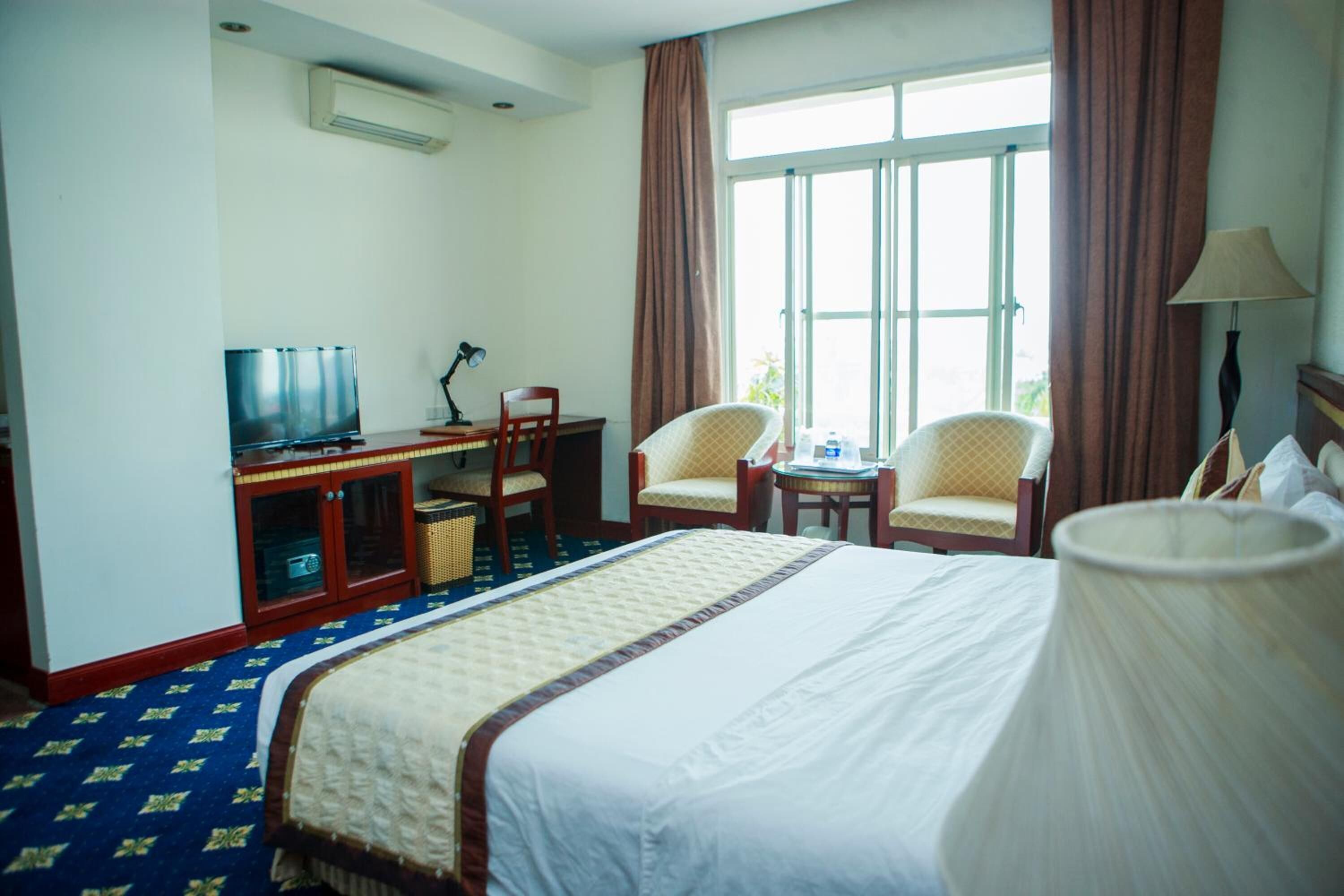Une chambre d’hôtel avec un grand lit, un bureau avec une télévision, deux chaises, une petite table avec une lampe et une fenêtre avec des rideaux.