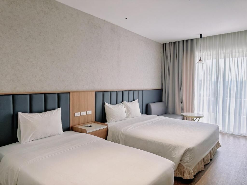 A hotel room with two beds, a headboard, a small table, and a window with curtains.