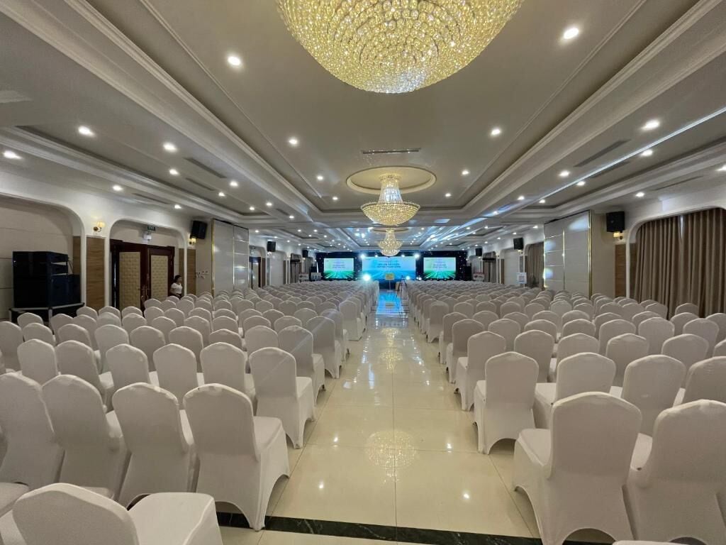 Un salón de conferencias con filas de sillas blancas, una lámpara de araña grande y varias pantallas al frente.