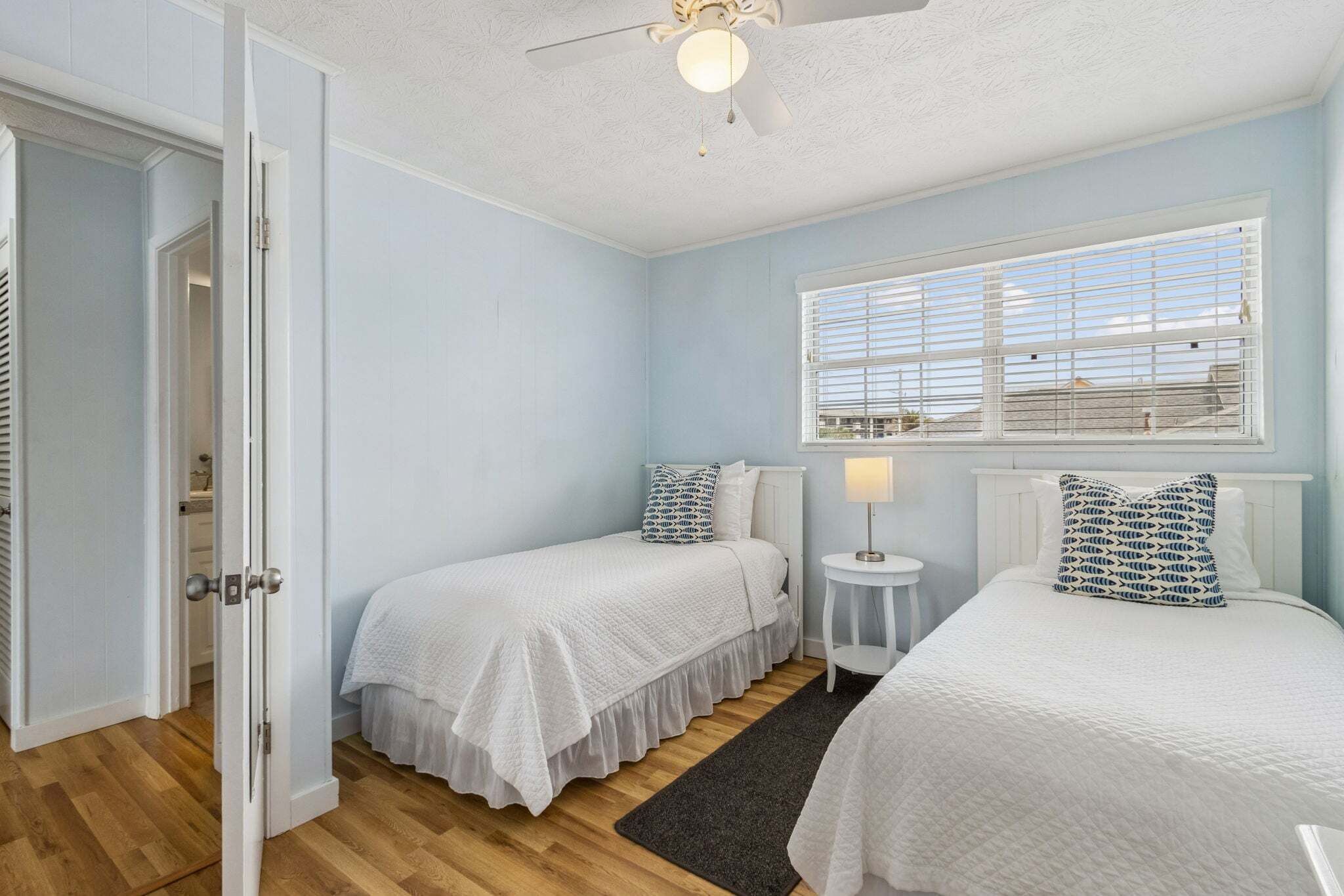 Une chambre avec deux lits, une fenêtre avec des stores, un ventilateur de plafond et une porte.