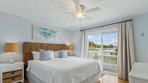Ein Schlafzimmer mit Bett, Deckenventilator, Nachttisch, Lampe, Fenster mit Vorhängen und Blick ins Grüne.
