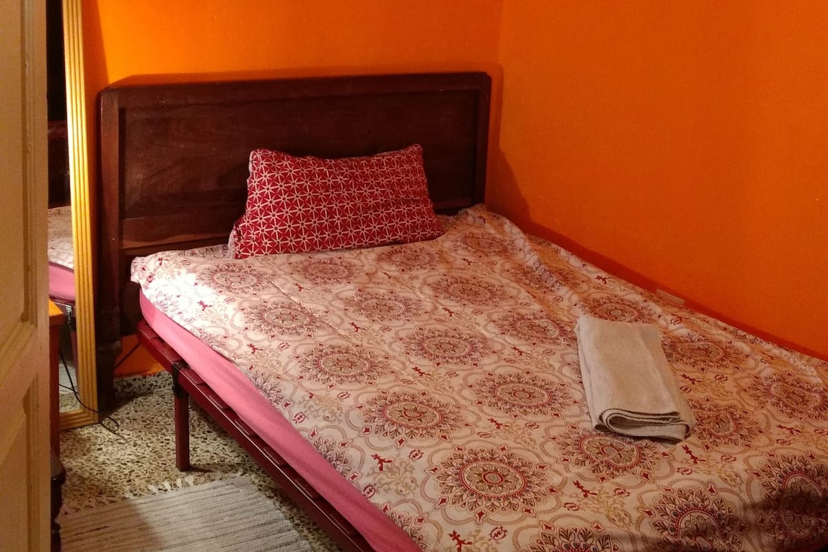A room with an orange wall, a wooden bed with a patterned bedspread, a red pillow, and a framed painting.